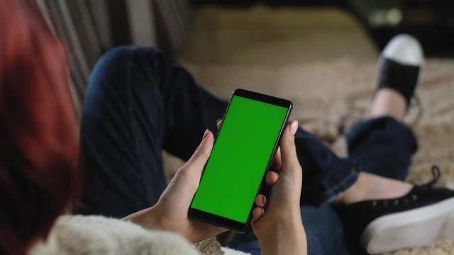 girl flips through photos with her fingers using her green screen phone. smartphone chromakey. woman in jeans is resting at home on couch and holding smartphone. green mockup to insert apps or images.
