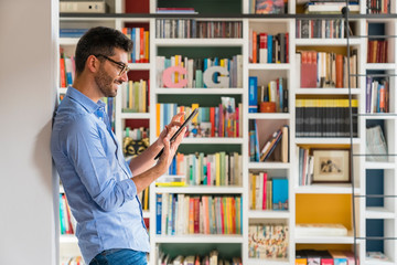 Smiling young man standing in front of bookshelves at home using digital tablet