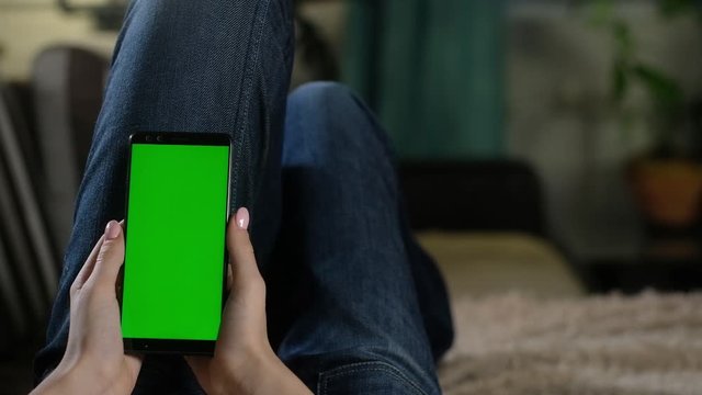 girl is resting at home on the couch and holding a smartphone with a green screen. green mockup to insert apps or images. Internet addiction. Gesture flipping photos with your fingers