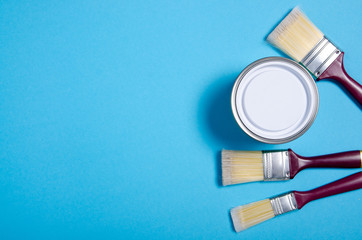 Open can with white paint and brush on blue background, top view. Space for text