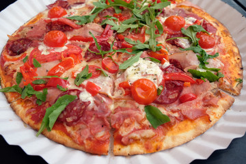 Pizza on a white paper plate. Delicious appetizing pizza cut into pieces with salami, mozzarella, red tomatoes and arugula. Fast food in disposable dishes.