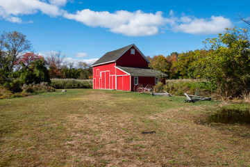 Fototapeta na wymiar Isolated red barn farmhouse in wooded tree area. Farmhouse surrounded by nature. Red barn with antique plow. 