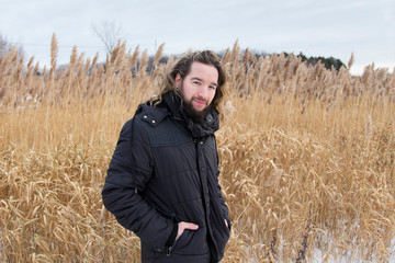 Naklejka na ściany i meble Medium horizontal view of young bearded man in dark jacket and long hair tied back turning and staring in a field during a early winter grey windy afternoon, Quebec City, Quebec, Canada
