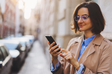 Side view of smiling beautiful adult woman in trench coat and eyeglasses using smartphone while standing beside traffic lane in daylight