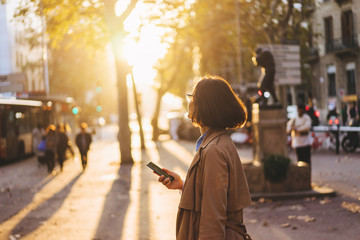 Girl enjoying sunset and chatting with friends via smartphone on the city streets - obrazy, fototapety, plakaty