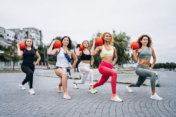 A group of sporty women are posing at camera, performing exercises with orange balls, on the street near the water. Active lifestyle