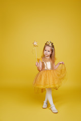 Joyful little girl with long hair in a tulle golden dress and princess crown holding a magic wand  on yellow background. Celebrating a colorful carnival for kids, expressing positive birthday