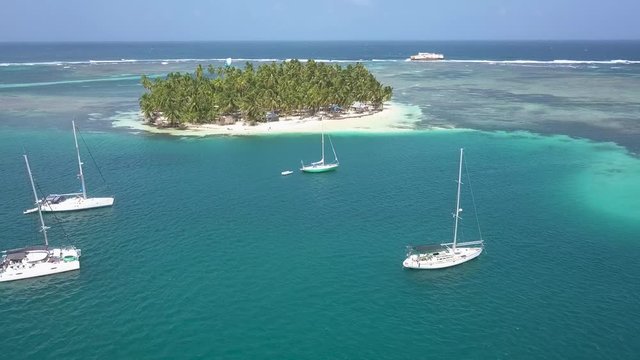 A flyover drone shot of some white sailing boats anchoring in a beautiful blue lagoon in front of a tropical island on a sunny day in San Blas, Panama.