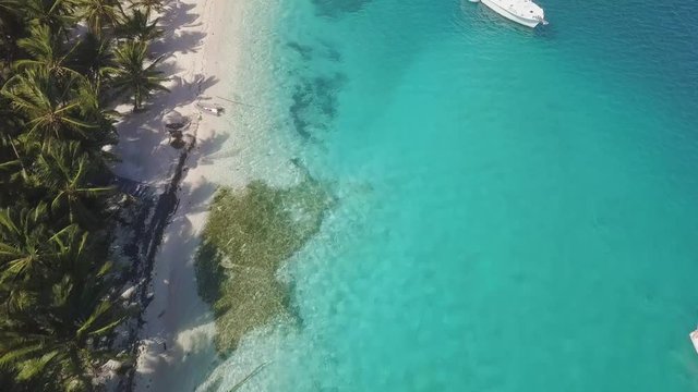 A nice drone shot flying over a big motor yacht anchoring in turquoise water next to the white sand beach of a tropical island overgrown with green palm trees on a sunny day in San Blas, Panama.