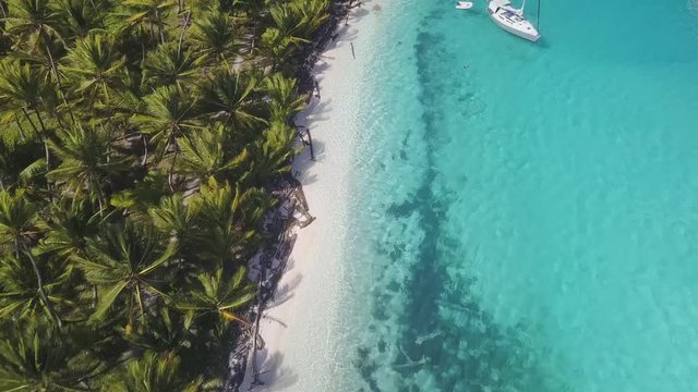 An amazing aerial shot flying over a sailboat anchoring in turquoise water next to the white sand beach of a tropical island overgrown with green palm trees on a sunny day in San Blas, Panama.