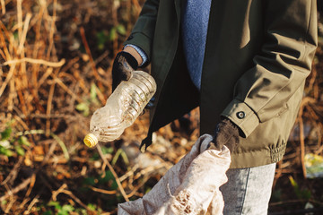Volunteer picking up dirty plastic bottles in park. Woman hand in glove picking up trash, collecting garbage in bag. Cleaning up nature from single use plastic