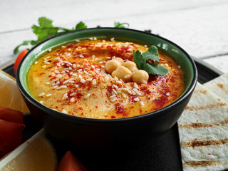 Bowl with traditional homemade hummus with olive oil and fresh vegetables. Slices of lemon and pita nearby on a plate. Useful natural snacks. Background of wooden white boards. Close-up
