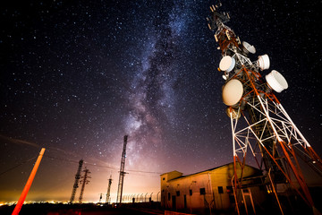 From below wonderful view of media antenna and silhouettes of electricity high voltage power towers against night city on horizon and gorgeous Milky Way in clear sky