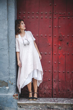 Thoughtful pensive serious dreaming stylish female in summer dress leaning on wall near ancient maroon door in town looking away