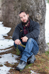 Full length vertical view of young bearded man in casual clothes and long hair tied back sitting relaxing at foot of evergreen tree during a early winter afternoon, Quebec City, Quebec, Canada