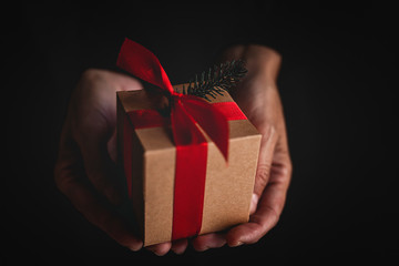 From above anonymous person demonstrating small gift box with red ribbon and conifer twig during Christmas celebration against black background