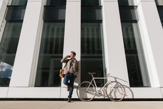 Low angle of serious entrepreneur with smartphone leaning on wall near a bicycle on modern building and looking away in downtown