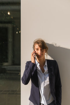 Optimistic businesswoman in dark jacket smiling and looking at camera while leaning on building wall on city street and answering phone call