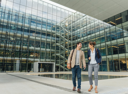 Full body delighted colleagues smiling and looking at each other while walking outside contemporary building after work