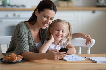 Happy mum and kid daughter using smartphone at kitchen table