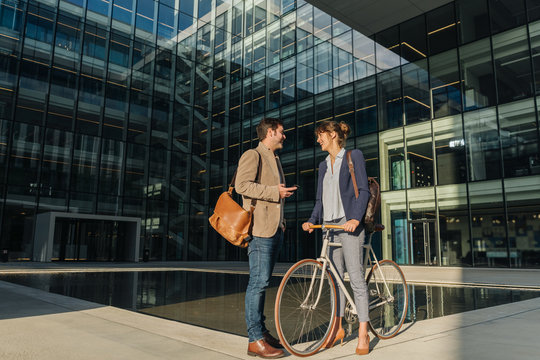 Cheerful man and woman with bicycle smiling and looking at each other while communicating outside office building on modern city street