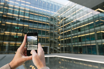 Crop adult male hands photographing transparent glass facade of contemporary high rise business center in downtown using smartphone