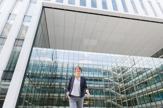 Low angle of delighted businesswoman smiling and walking from walk outside contemporary office building with glass walls