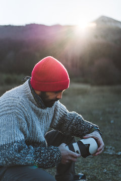 Side view of bearded male photographer in red knitted cap and grey sweater grabbing a professional camera in a backpack while in mountains