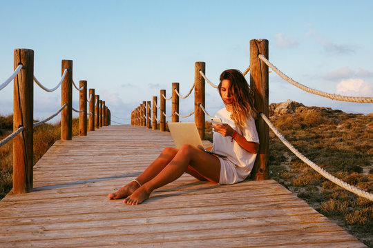 Concentrated female freelancer on vacation in white shirt sitting on empty bridge and working with laptop and using mobile phone on blue sky background