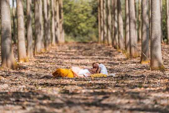 Side view of happy barefooted pregnant woman and little girl hugging and resting on ground while spending time together in autumn forest glade during sunny day