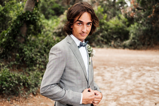 Young groom in stylish gray wedding suit going on empty road with green trees on sides