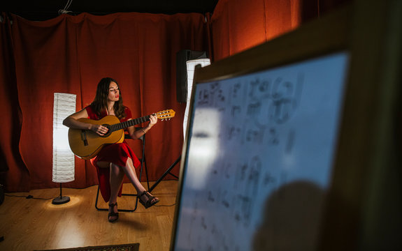 Charming artistic woman in red dress performing song playing on guitar in stage with warm light in Spain