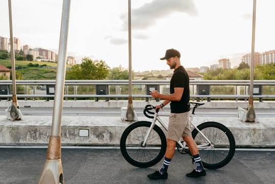 Side view of young casual bearded male cyclist in black cap using phone while walking with bike on road with city buildings on background