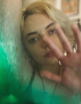 Through glass pensive young blond freckled female with brown eyes looking at camera and dreaming while touching transparent surface with green transition