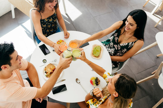 From above multi ethnic young people clinking glasses of fresh fruit beverages and proposing toast while sitting at table with healthy snacks in cafe