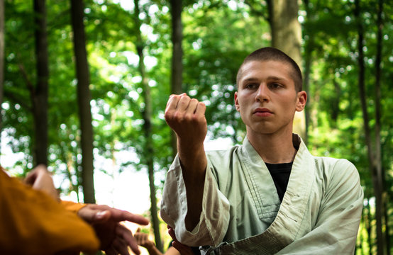 Serious bald man in uniform raising arm and looking away while practicing martial arts on blurred background of gazebo and green bushes in garden