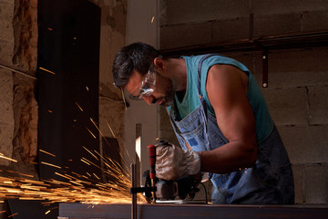 Side view of workman in protective glasses and gloves cutting metal with grinder with flying sparks while working in workshop