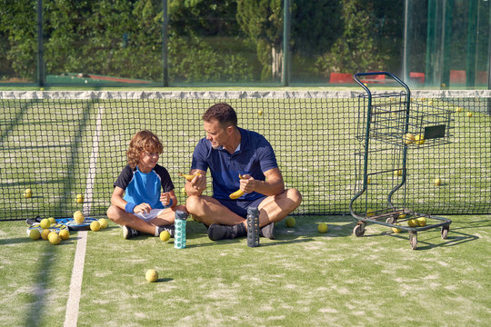 Cheerful adult man and boy smiling and eating bananas while sitting crossed legged near net on tennis court and resting
