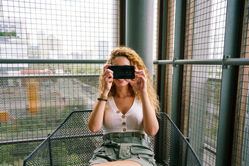 Smiling curly woman chilling in modern waiting area sitting in metal chair and taking photo with smartphone
