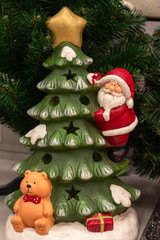 a compositional Christmas toy under the tree, Santa Claus with gifts climbs on the holiday tree, and underneath is a teddy bear with a cap. The toy symbolizes the New Year. Vertical photo