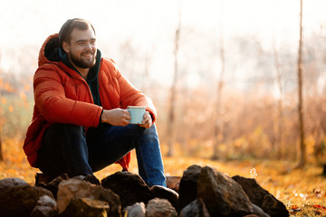 Style man in down jacket with cup of coffee sitting near bonfire