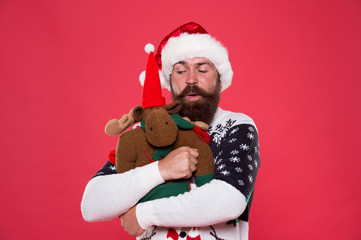 Christmas prep concept. Hipster bearded face winter sweater. Winter holiday. Soft toy. Hug and love. Buy gifts. Winter sale. Winter feast. Reindeer. Man santa hat hold christmas gift red background