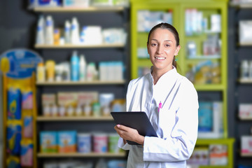 Beautiful young pharmacist standing next to medicine shelves, holding tablet