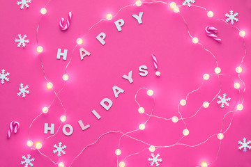 New Year or Christmas pattern flat lay top view background with text "Happy holidays". Paper snowflakes and garland of festive lights on pink background. Trendy glowing monochrome toned background.