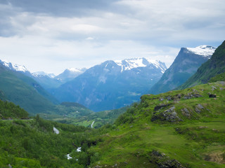 Fototapeta na wymiar Mountain landscape with snow on the peaks near Geiranger, Norway and a beautiful green valley with a river and winding highway up to viewpoint on a cloudy day.