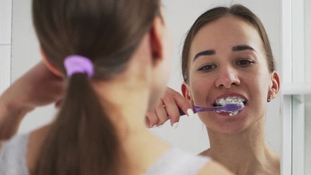 Oral hygiene for braces. Teen girl cleaning and brushing teeth with clear ceramic braces using special brushing tools. Toothpaste. There are mirrors in the bathroom. Close-up.