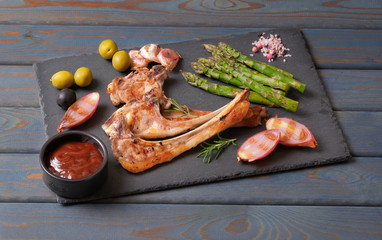 Baked ribs grilled, roasted lamb ribs with olives, asparagus, onions and garlic on natural black stone plate. Organic grilled mutton meat chops with vegetables and sauce on rustic wooden background.