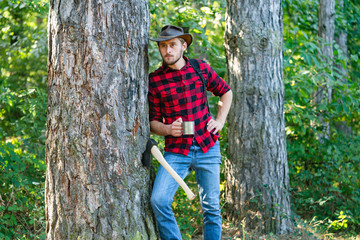 Naklejka na ściany i meble Harvest of timber. Bearded man with axe concept. Deforestation. Handsome Woodworkers lumberjack plaid shirt holding the axe on green nature background.