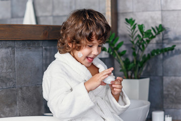 Handsome little funny boy with dark hair in white dressing gown taking skin care cream on finger and putting the stain on his face in the bathroom