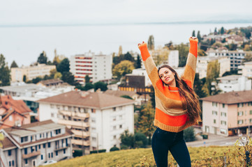 Outdoor portrait of beautiful young woman wearing warm pullover, posing on city background, arms wide open